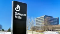 A prominent black sign featuring the white General Mills logo and text 'General Mills' is centered on the left, set against a bright blue sky. To the right, a multi-story modern office building with many windows stands, surrounded by bare trees and snow-covered ground under the clear sky.
