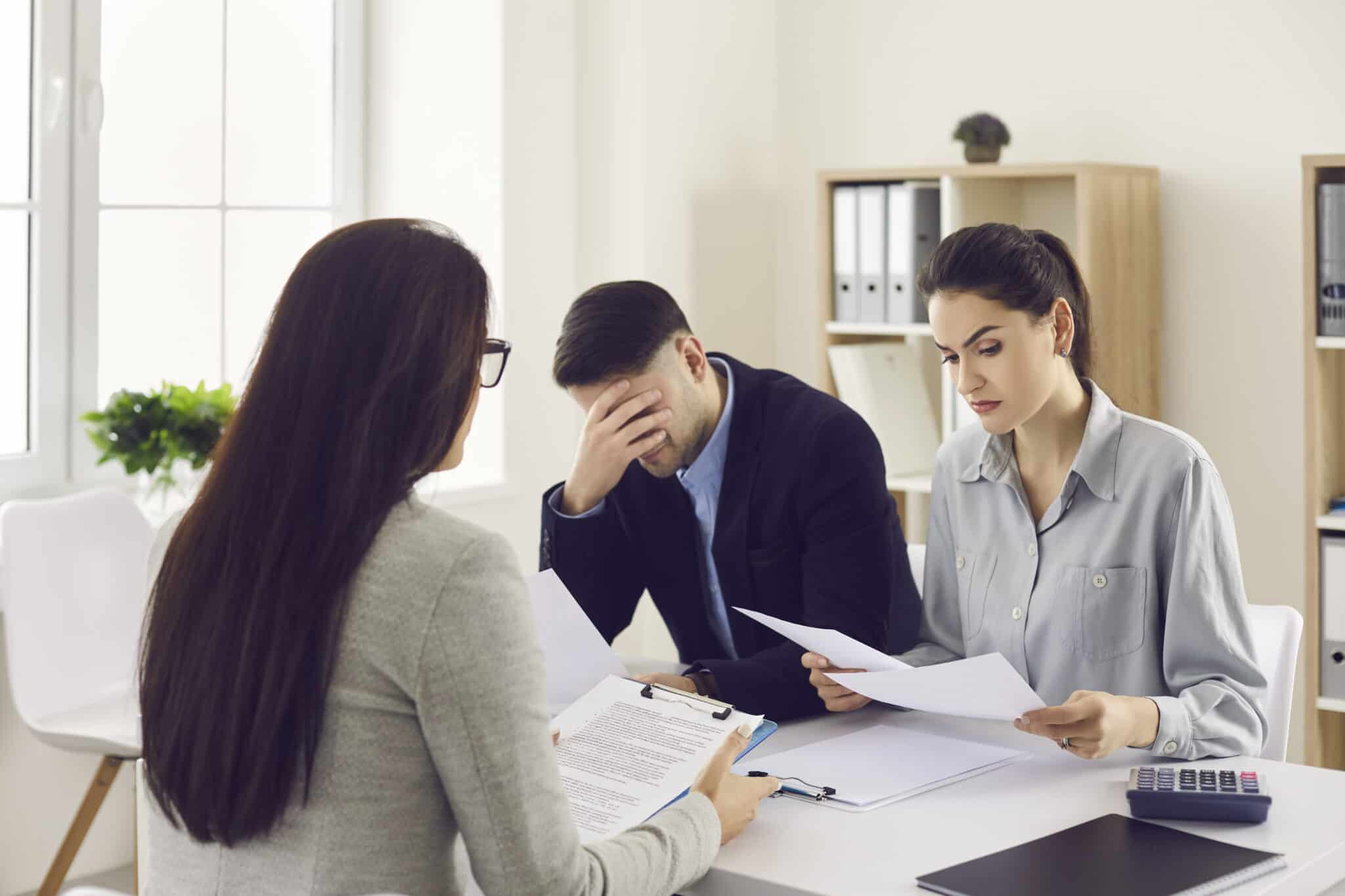 Three people are seated at a white table in an office. On the left, a woman with long dark hair and glasses, seen from behind, holds a clipboard with papers. In