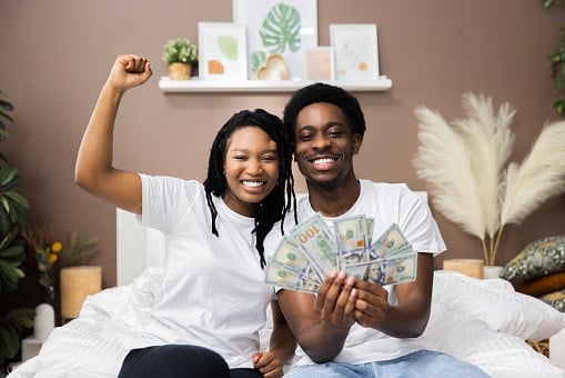 Smiled young African American dark skinned couple man and woman wearing casual white t-shirt smiling raising fists holding a lot of money cash in hands celebrating happi lottery winners