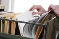 A close-up view of two hands, one on the left and one on the right, actively sifting through a gray metal filing cabinet filled with numerous light brown and tan hanging file folders. White papers are visible within the folders, some with text. The background is bright and out of focus.