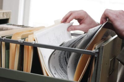 A close-up view of two hands, one on the left and one on the right, actively sifting through a gray metal filing cabinet filled with numerous light brown and tan hanging file folders. White papers are visible within the folders, some with text. The background is bright and out of focus.