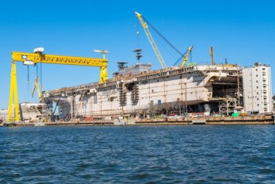 A large, unfinished naval ship, mostly white, sits in a shipyard dry dock. A massive yellow gantry crane with 'Ingalls Shipbuilding' written on its side spans over the vessel. Numerous smaller cranes, scaffolding, and buildings surround the ship, indicating active construction. Blue water is in the foreground, and a clear blue sky is above.