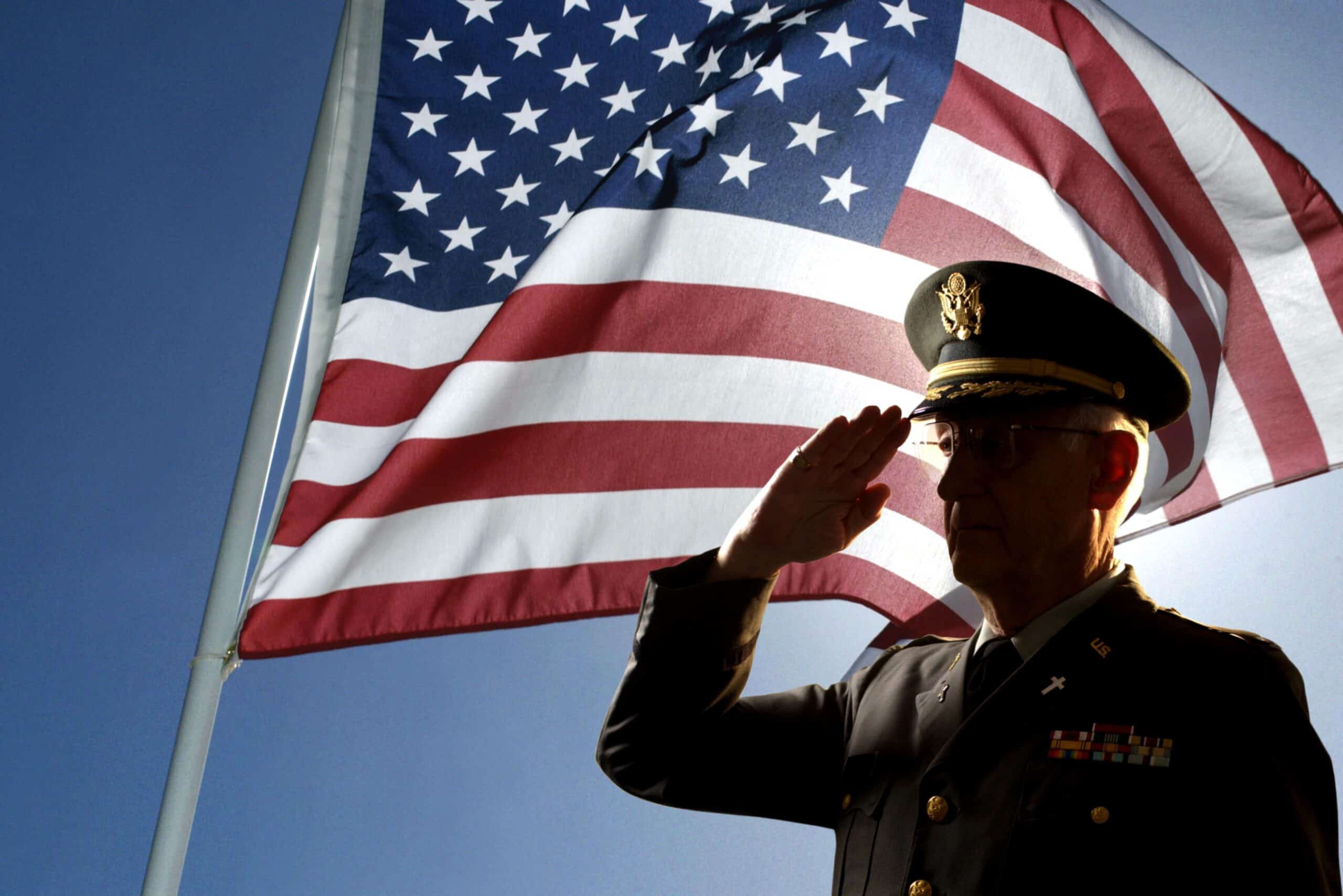 Army: Colonel | Silhouette of veteran US Army Colonel Chaplain wearing hat and saluting with an American flag flying behind him.
