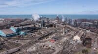 An aerial, wide shot of a vast industrial steel manufacturing plant located on the shore of a large body of water. Numerous gray and blue industrial buildings, some with smoking chimneys, are interconnected by extensive rail lines and conveyor belts. Piles of dark aggregate material are scattered throughout the facility. The sky above is light blue with scattered white clouds.