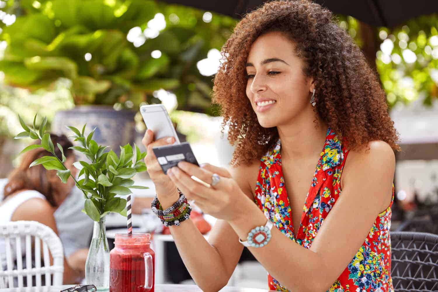 Shot of beautiful glad young female with Afro hairdo, types number of credit card on smart phone, makes purchase online or checks bank account, recreats in outdoor cozy cafe with fresh cocktail