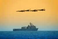 Four dark-silhouetted fighter jets fly in a precise formation against a vibrant orange and yellow sky. Below them, a large gray naval ship, possibly a warship or amphibious vessel, sails on choppy deep blue ocean water. An American flag flies prominently from the ship's mast, adding to the patriotic and powerful scene.