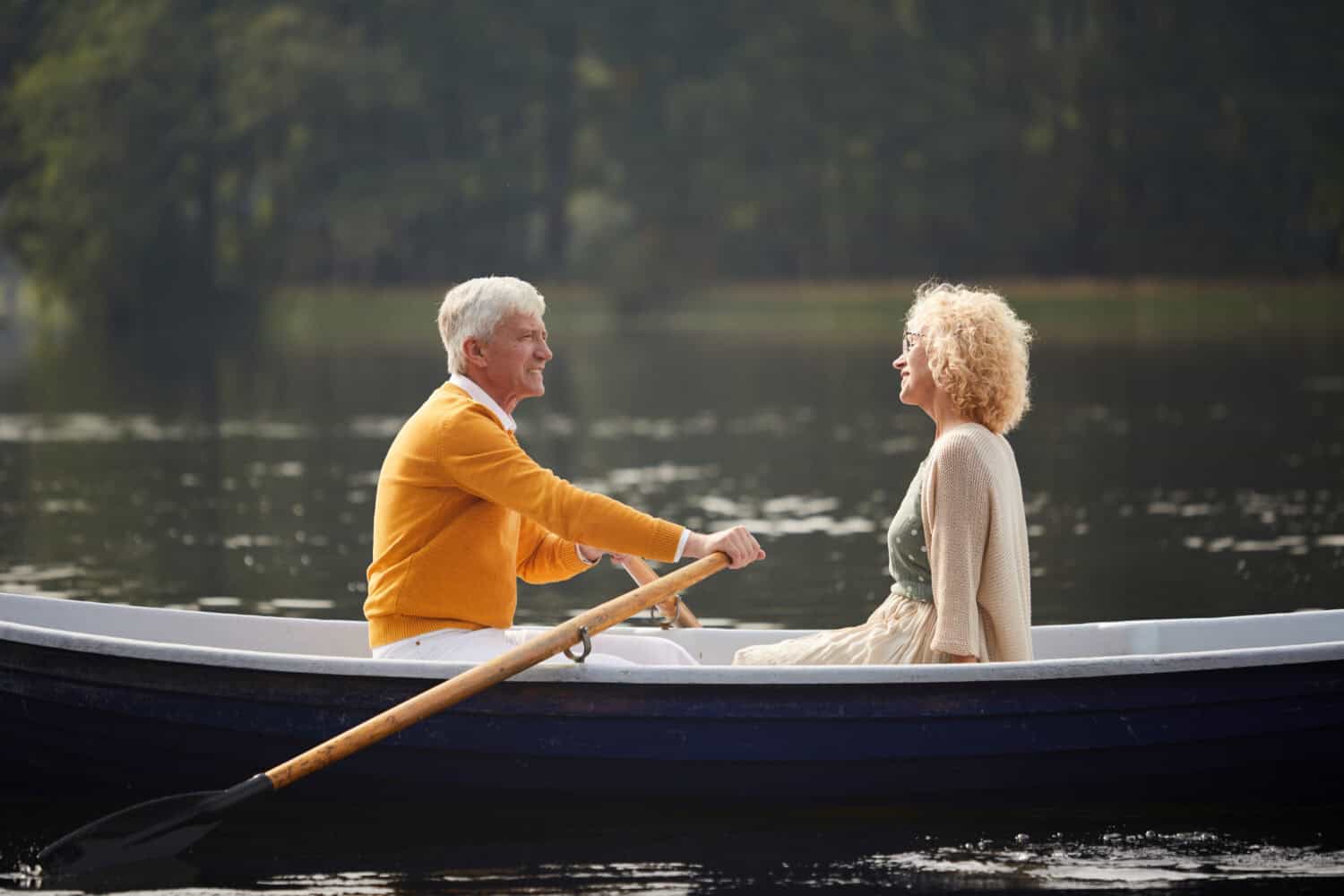 Happy carefree senior couple riding boat on lake and talking, handsome elderly man rowing oars