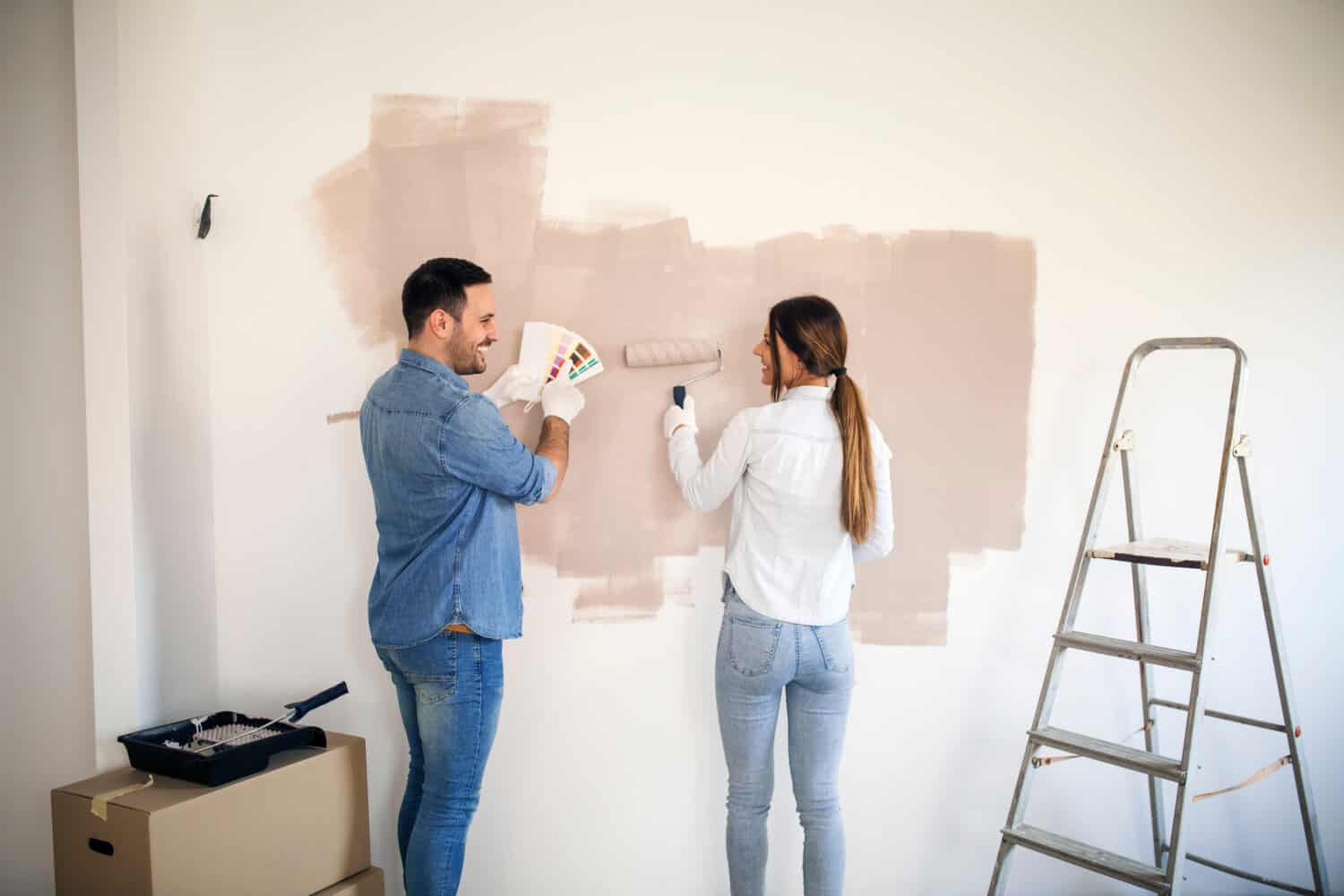 Couple renovating their apartment while man holding color palette and woman painting walls with roller.