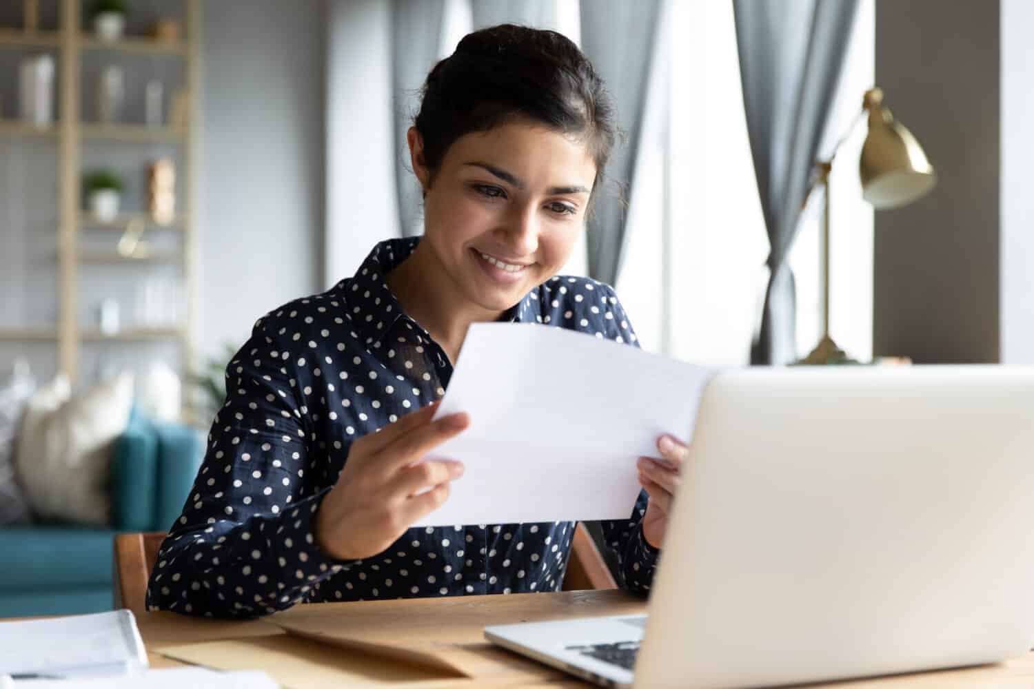 Smiling young indian woman holding paper bill letter doing paperwork bill reading good news check post mail sit at home table, happy lady customer receive bank receipt sheet tax refund notification