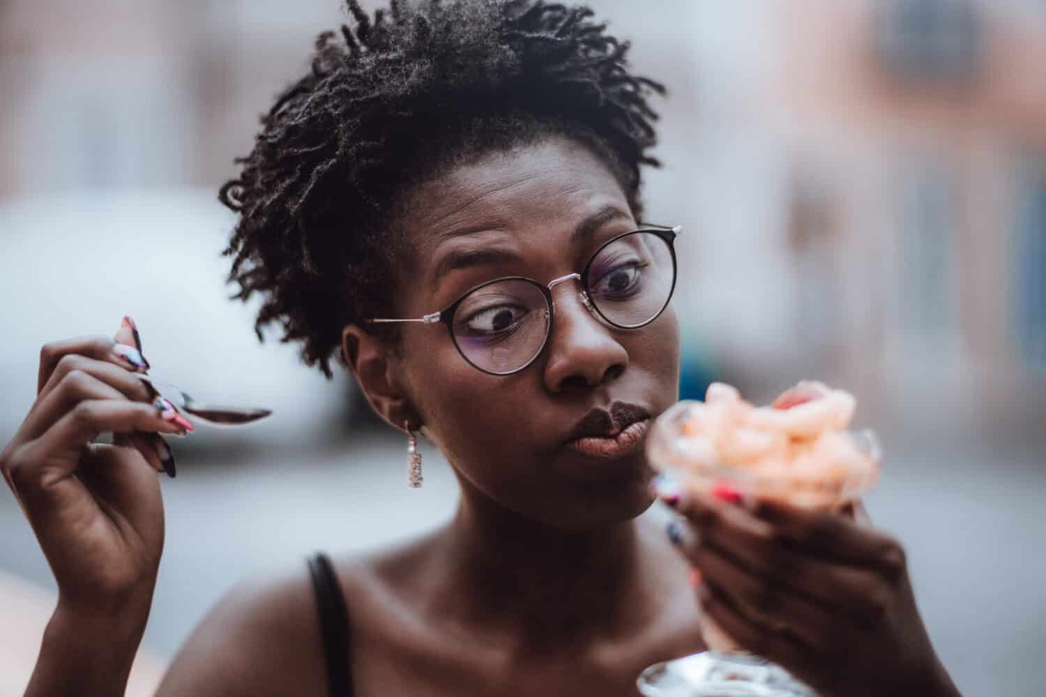 Young charming funny African female in eyeglasses and with a nail-art is surprised at the taste of the dessert that she just started to eat and tasted while sitting in an outdoor cafe or a restaurant