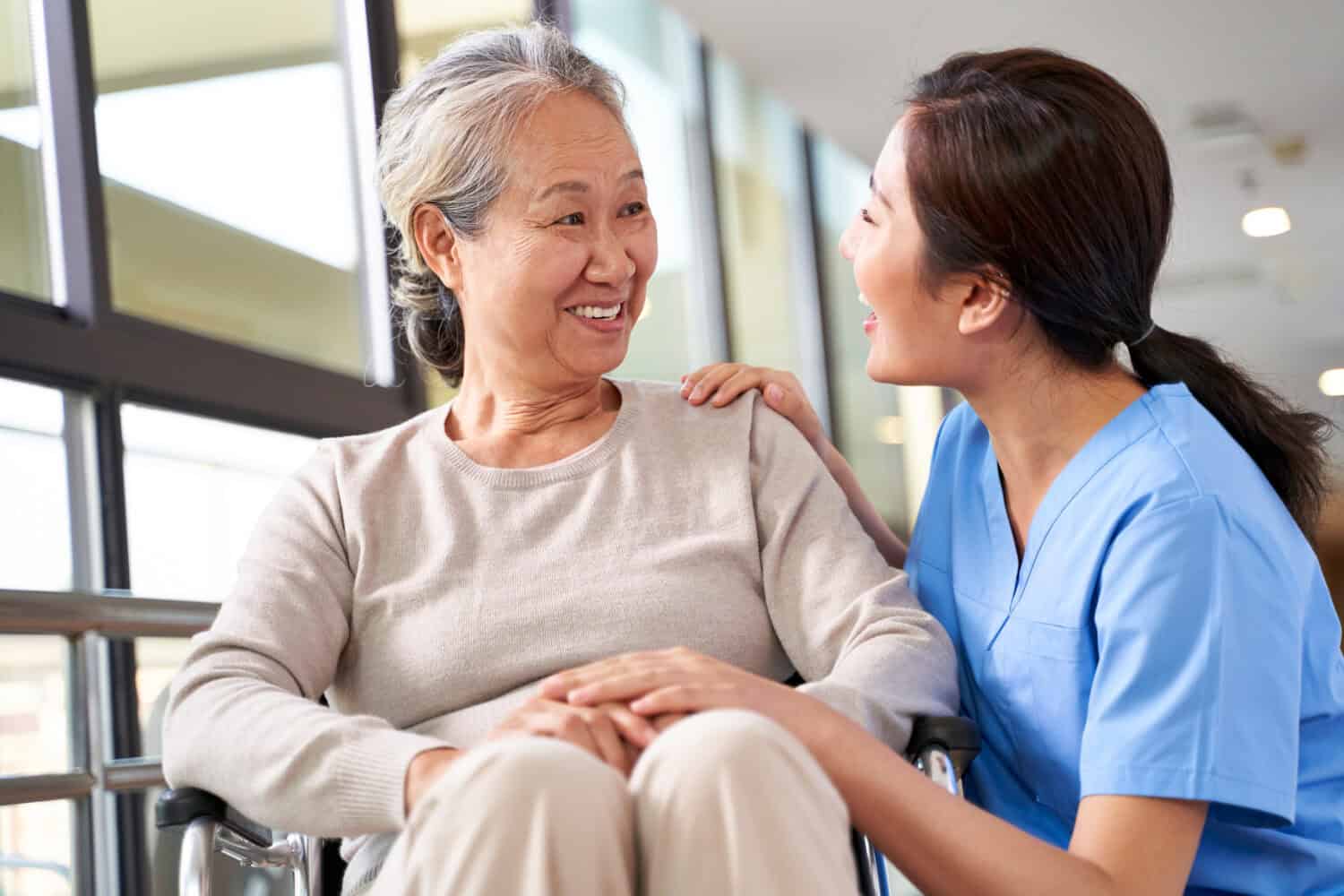 friendly caregiver of nursing home talking to asian senior woman in hallway