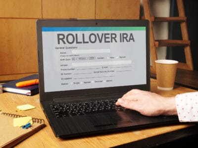 A person's hand with a white shirt sleeve types on the black keyboard of an open laptop. The laptop screen displays a 'ROLLOVER IRA' form with the title 'General Questions'. Pre-filled fields show 'Birth Date: 01 January 2000'. The laptop is on a light wooden desk, with notebooks and a pen visible in the foreground.