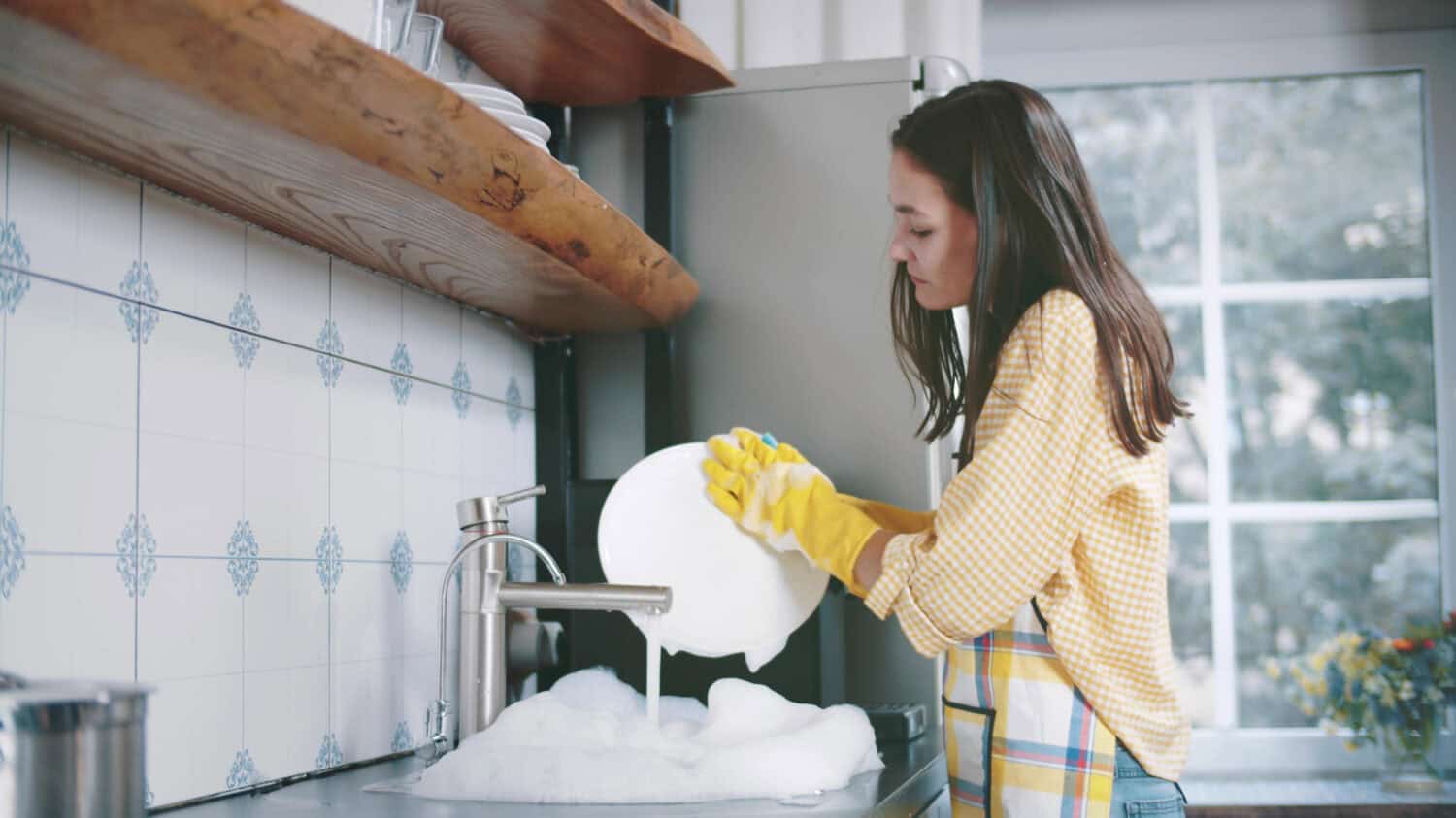 Happy attractive woman in apron and gloves washing dishes at home kitchen. Side view of pretty young housewife doing household. Beautiful female wash plates
