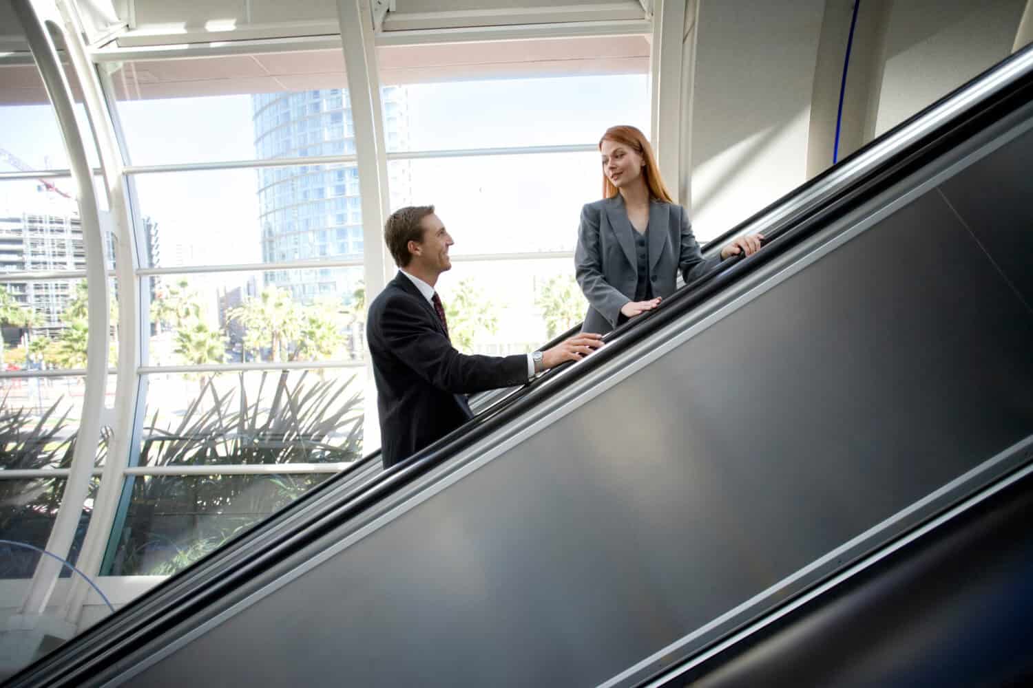 Horizontal profile shot of a businessman and businesswoman smiling at each other on an escalator going up in a building.