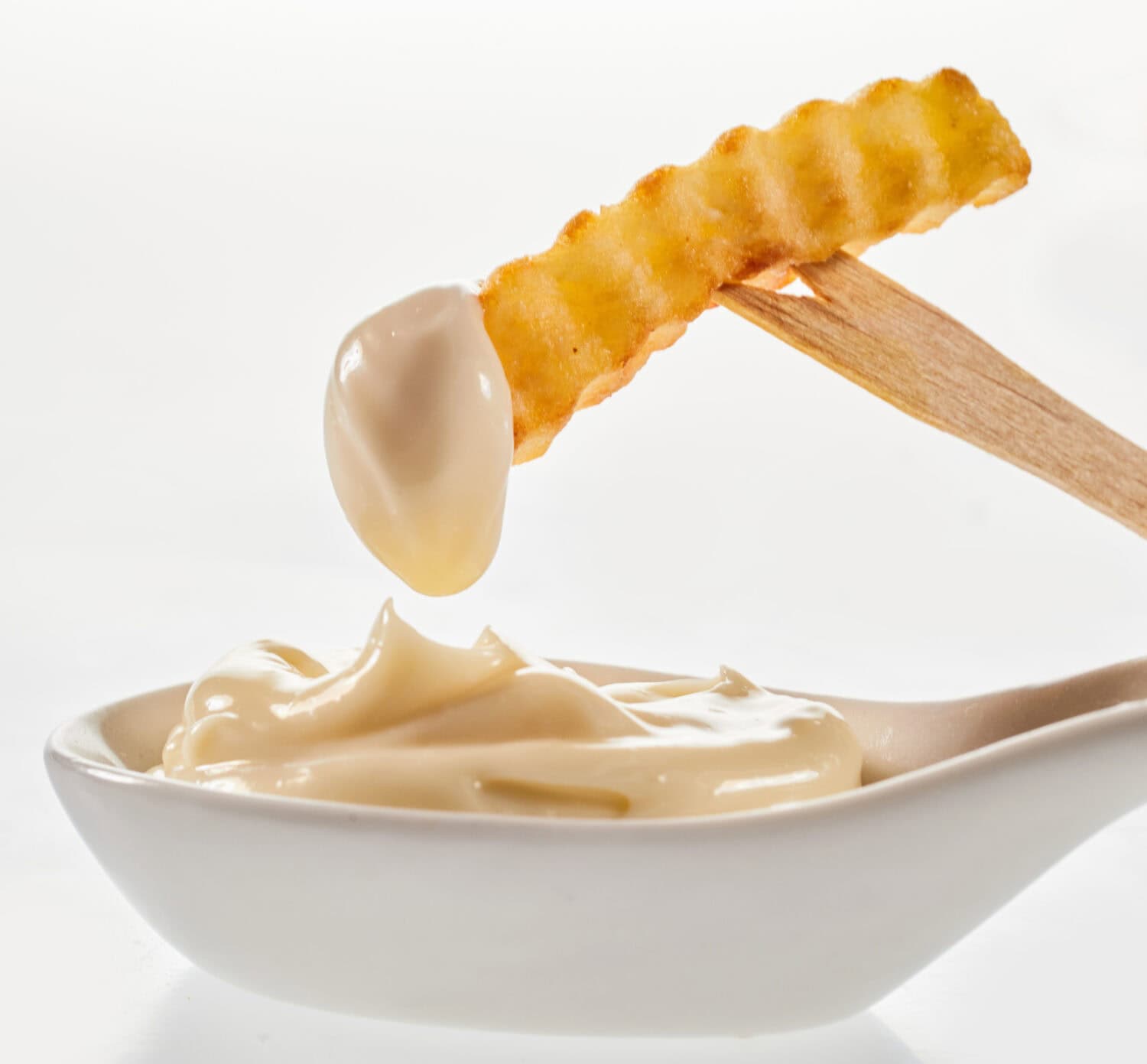 Appetizing French fried potato stick with ribbed surface on wooden fork over ceramic spoon with creamy mayo on white background