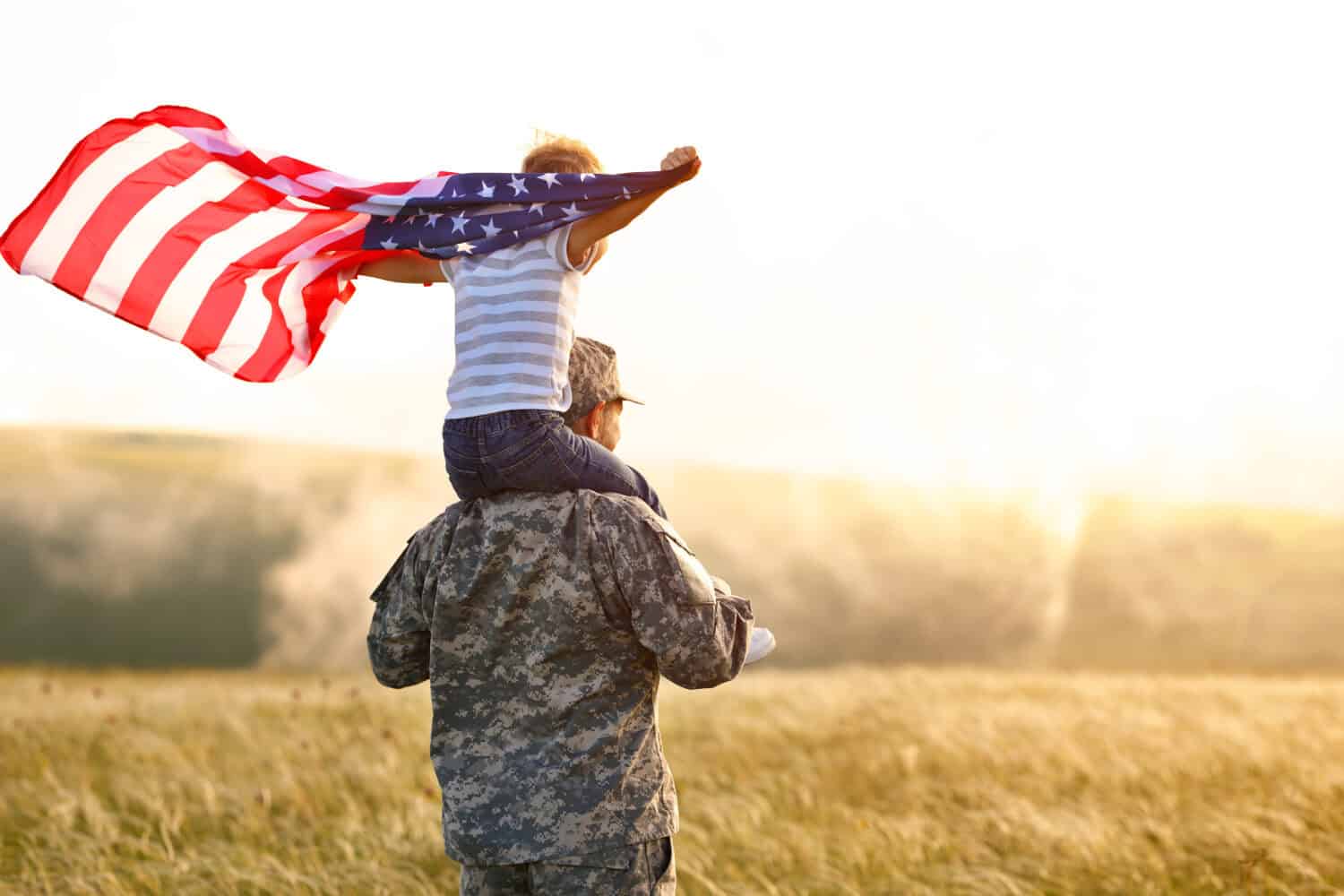 Rear view of military man father carrying happy little son with american flag on shoulders and enjoying amazing summer nature view on sunny day, happy male soldier dad reunited with son after US army by Evgeny Atamanenko