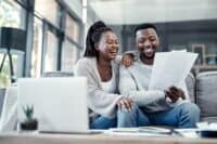 A joyful Black couple sits closely on a gray sofa in a modern living room. The woman, with braided hair in a high ponytail, rests her arm on the man's shoulder as they both laugh while he holds and looks at white papers. A white laptop sits on a table in the foreground, slightly out of focus.