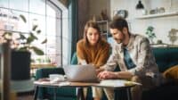 Stressful Accounting at Home: Couple Using Laptop Computer, Sitting on Sofa in Apartment. Young Family Filling Tax Forms, Mortgage Documents, Bills, Checks, Balances, Invoices are in Order