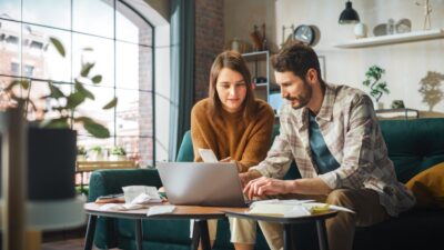 Stressful Accounting at Home: Couple Using Laptop Computer, Sitting on Sofa in Apartment. Young Family Filling Tax Forms, Mortgage Documents, Bills, Checks, Balances, Invoices are in Order