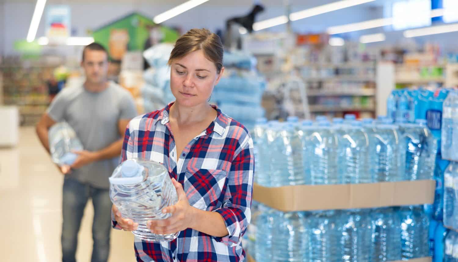 Interested young woman choosing bottled still water in supermarket, reading label on plastic bottle..