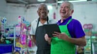 Two smiling older men, both wearing aprons, stand in what appears to be a retail store. The man on the right, with gray hair and a green apron over a purple shirt, holds a black tablet and laughs heartily. The man on the left, with dark skin and a black apron over a white collared shirt, also laughs, looking towards the viewer. Colorful merchandise shelves are blurred in the background.