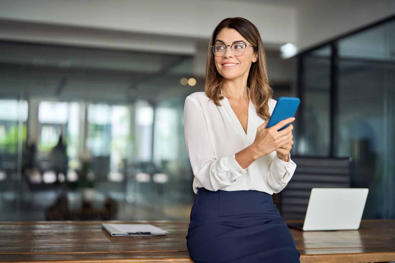 Happy mature business woman executive holding cell phone looking away in office. Smiling mid aged 40s professional businesswoman manager entrepreneur using cellphone working on smartphone. Copy space