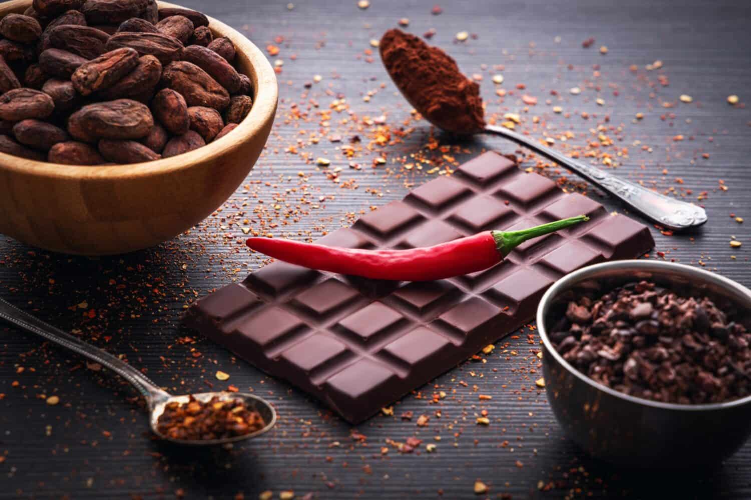 Dark chocolate bar, red hot chilli pepper cayenne, dry hot chili spices, cocoa beans nibs powder, food tasty design on black wooden background