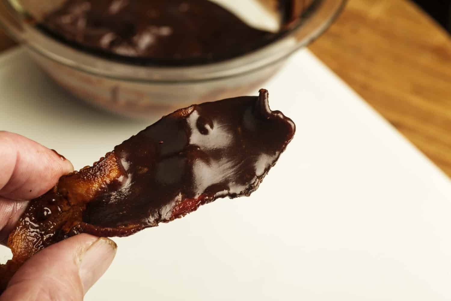 Cooked bacon slice covered in dark chocolate ganache with glass chocolate-filled bowl in the background