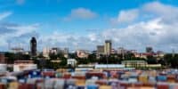 An aerial tilt-shift photograph of a city skyline under a bright blue sky with scattered white and grey clouds. In the blurred foreground, many colorful shipping containers are stacked. The mid-ground reveals a dense urban area with diverse buildings, including modern high-rises, a historic church with a prominent spire, and residential structures. Green trees are visible between the buildings, adding natural elements to the cityscape.