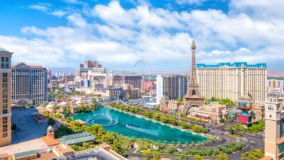 A high-angle daytime view of the Las Vegas Strip. In the foreground, a large turquoise lake with elaborate fountains spans the center, surrounded by palm trees and hotel properties. To the right, a replica of the Eiffel Tower stands prominently next to a large building with blue rooftops. Further back, numerous skyscrapers and resorts line the street, with the High Roller observation wheel visible in the distance under a bright blue sky with white clouds.