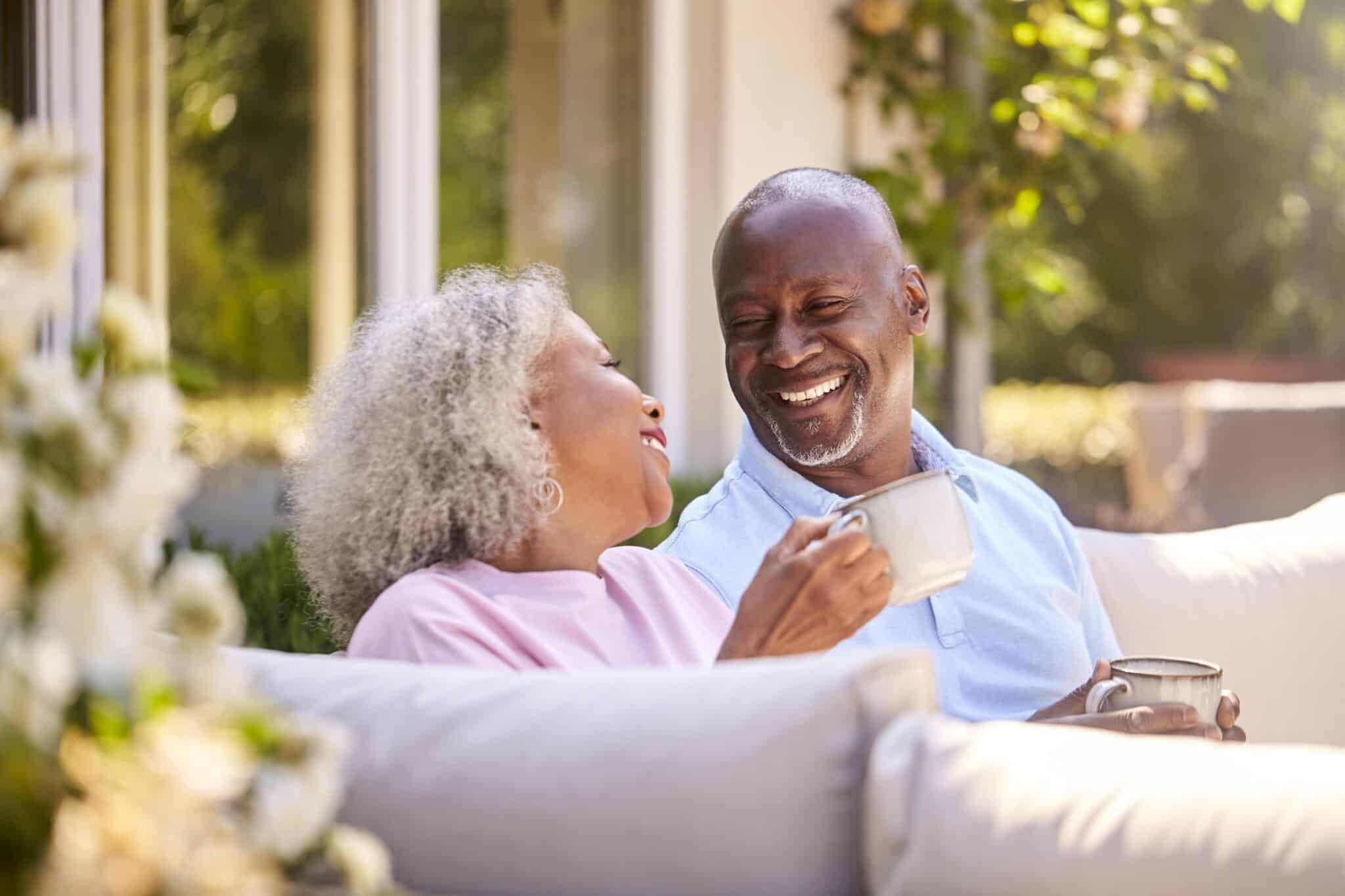 An older Black couple sits closely on an outdoor couch, both smiling brightly and looking at each other. The woman on the left, with grey curly hair, holds a wh