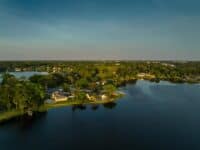 An aerial view showcases a residential area bordering a dark blue lake under a clear, gradient sky of blue and light grey. Numerous houses with varied rooflines are visible, many featuring private swimming pools, and surrounded by dense, vibrant green trees. The composition emphasizes the waterfront properties and the suburban landscape.