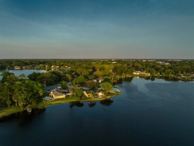 An aerial view showcases a residential area bordering a dark blue lake under a clear, gradient sky of blue and light grey. Numerous houses with varied rooflines are visible, many featuring private swimming pools, and surrounded by dense, vibrant green trees. The composition emphasizes the waterfront properties and the suburban landscape.