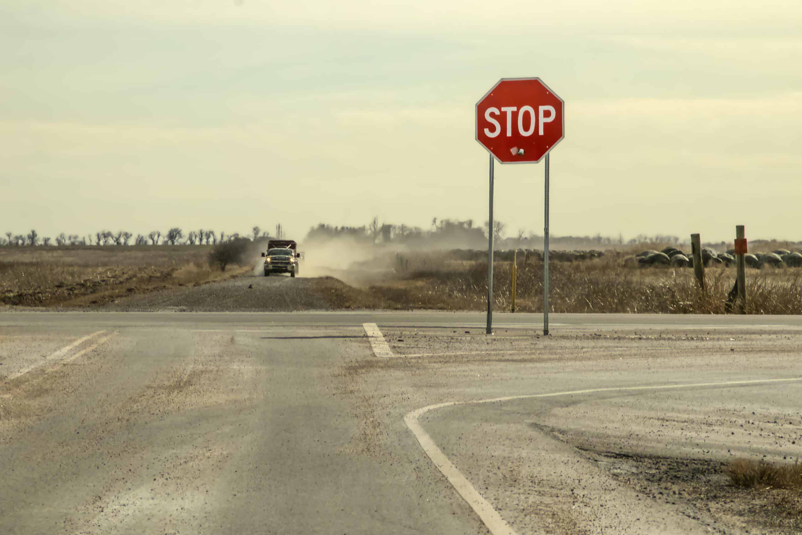 Oklahoma gun | Country Roads - Stop sign with large bullet hole stands by crossroads with pickup truck with trailer driving toward it in a cloud of dust