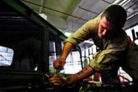 A young man with short hair and a tan work uniform leans over a vehicle engine or complex machinery, his hands holding tools and manipulating components. He is intently focused on his work, illuminated by overhead lighting in what appears to be a garage or workshop. A tattoo of 'E=MC^2' is visible on his right forearm, and the composition captures the detail of his concentrated effort.