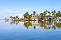 A panoramic view of several colorful houses nestled among dense green palm trees along a tranquil blue waterfront. The sky is bright blue with a few wispy white clouds. The water perfectly reflects the houses and trees, creating a mirror-like surface. On the far left, a thatched-roof structure extends into the water. Boats are docked near some of the homes.