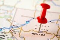 A close-up, shallow depth of field shot of a red pushpin stuck into a physical map. The pushpin is prominently centered on the state of Nevada, which is clearly labeled in capital letters. Other visible states and cities include Reno, Carson City, and Sacramento, along with several highway markers like I-80, US-50, and US-395. The map has a textured, paper-like appearance.