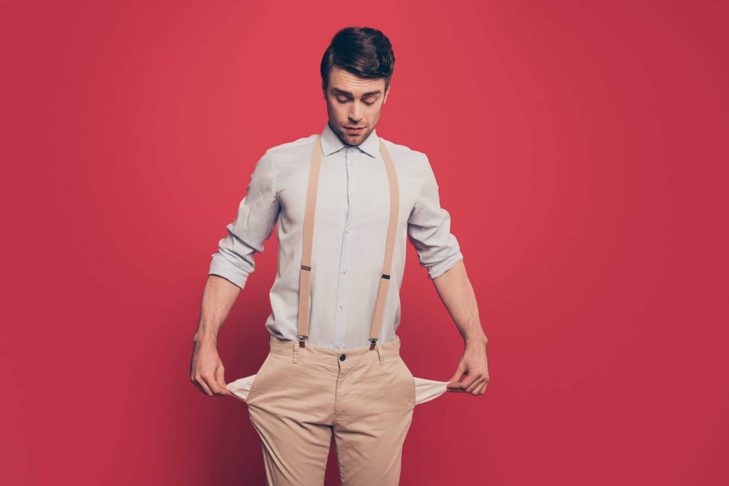 Professional, cunning magician, illusionist, gambler in casual outfit, showing two empty pockets out, standing over red background, looking down
