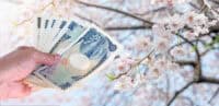 A close-up shot of a hand holding a fan of Japanese Yen banknotes, including 1000 and 10000 yen denominations. The background is softly blurred, revealing light pink and white cherry blossoms (sakura) on tree branches under a bright sky.