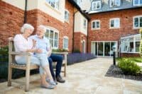 A happy elderly couple sits on a wooden slatted bench outdoors. The woman, with white hair and wearing a pink top and light grey pants, faces the man, who has a white beard, a light blue collared shirt, and dark blue pants. They are smiling at each other. In the background is a large brick building with multiple white-framed windows, a paved stone patio, and a border of purple lavender plants along the path.