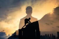 A double exposure image featuring the dark silhouette of a person in a business suit facing away from the viewer, overlaid with a dynamic silhouette of a person climbing a rocky incline. The background is a vivid sunset sky with hues of orange, yellow, and deep blue, revealing distant silhouetted city buildings and a natural hill.