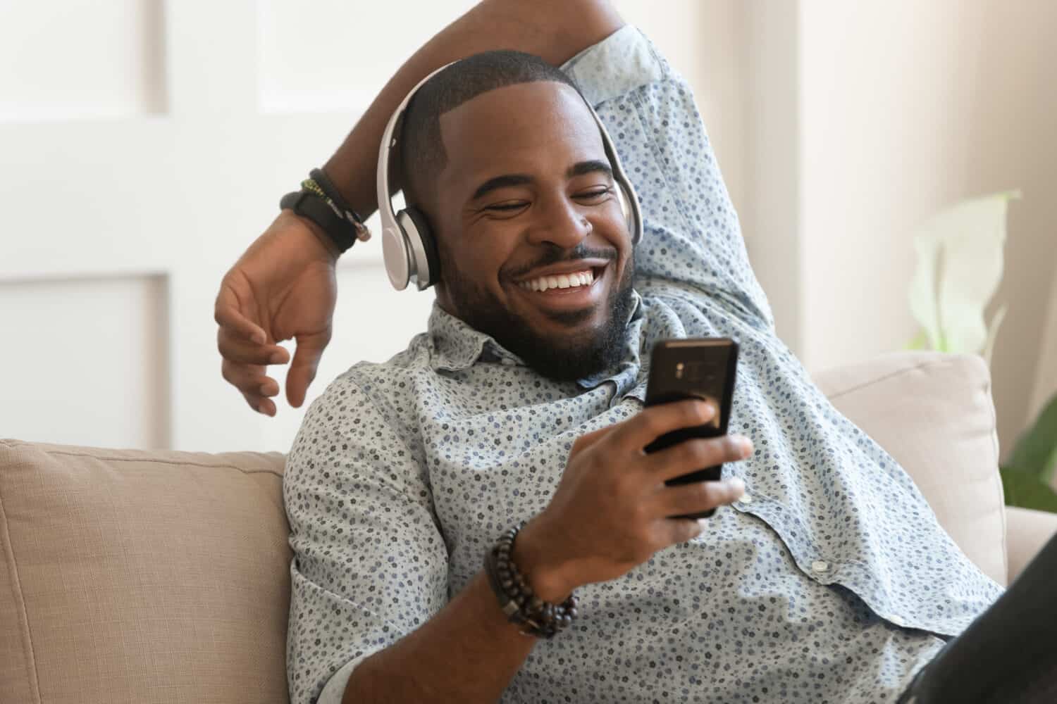 Smiling young african american man wearing headphones listen to mobile music playing in smartphone app, happy black guy relaxing holding using phone enjoy favorite songs relaxing sit on sofa at home