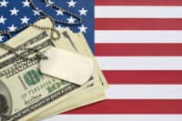 A silver-colored military dog tag is prominently displayed on top of a stack of US hundred-dollar bills. Both items are laid over a textured American flag, with the blue field and white stars visible on the left and the red and white stripes on the right side of the frame.