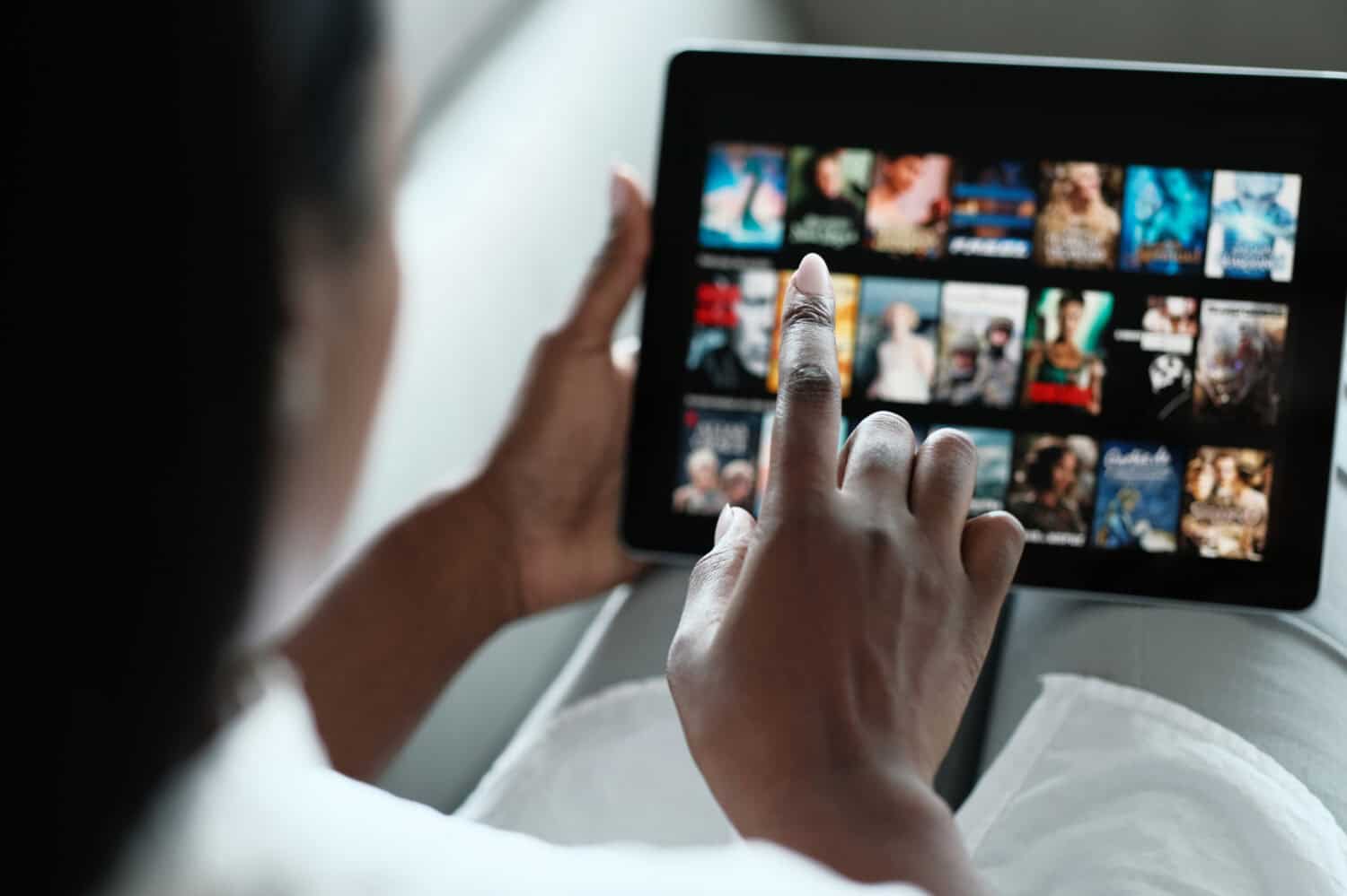 African American Woman Lying Down On Sofa At Home, Choosing Movie On Internet Streaming Service. Over The Shoulders.