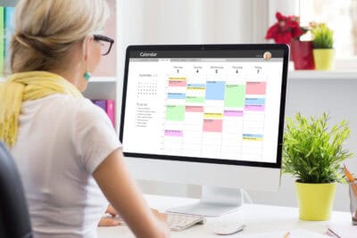 A woman with blonde hair and glasses, wearing a white shirt and yellow scarf, sits at a desk typing on a white keyboard. She is looking at a large computer monitor displaying a calendar application with multiple colored appointments for September 3-7. A green potted plant and a pen holder are visible on the desk to the right.