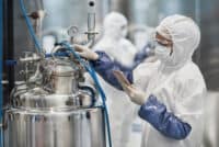 A person in a white cleanroom suit, face mask, safety glasses, and blue over-sleeves stands next to a large, shiny, stainless steel bioreactor. The person is wearing white gloves and holding a tablet in their left hand, while their right hand is near blue tubing connected to the top of the vessel. A pressure gauge is visible on the bioreactor, and another blurred figure in a cleanroom suit is in the background.