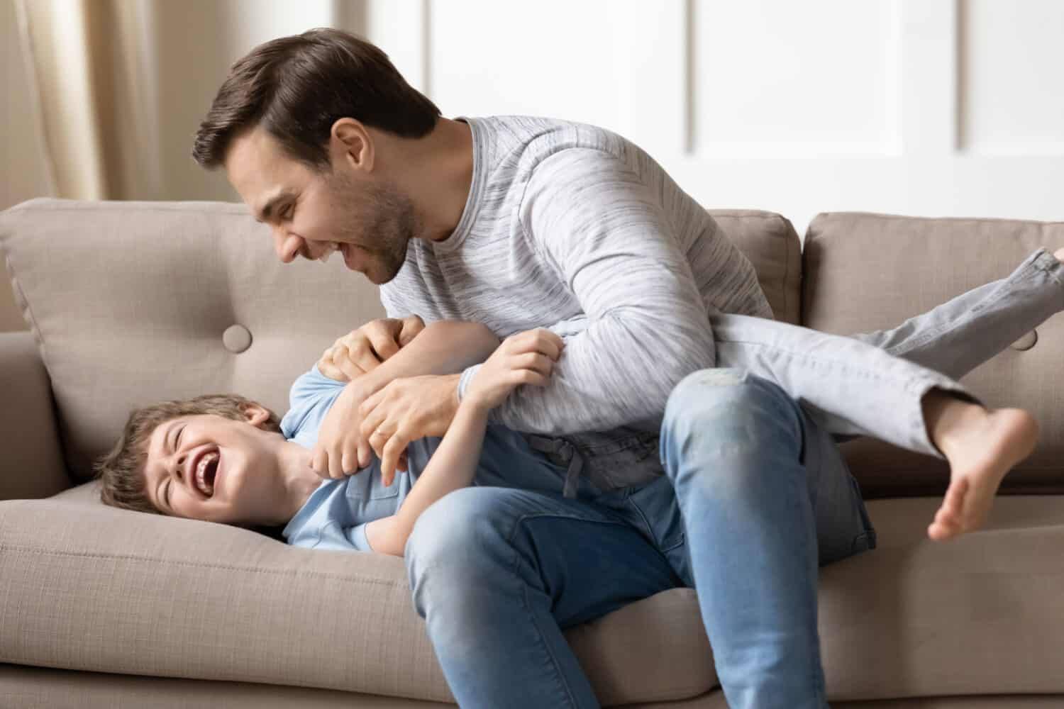 Playful young Caucasian father play with excited smiling small 8s son in living room at home. Overjoyed dad have fun with little preschooler boy child, tickle giggle, enjoy family weekend together.
