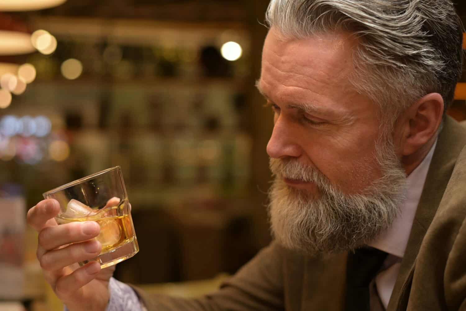 The gray-haired man looks at the whiskey glass in his hand. Adult man drinking in a restaurant