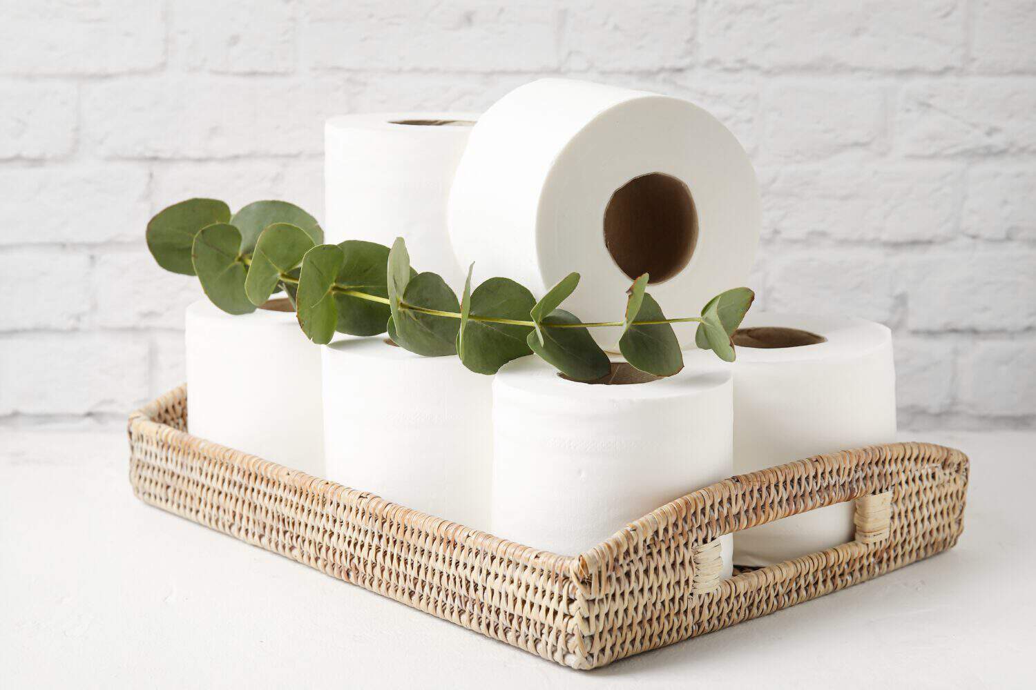 Basket with rolls of toilet paper and eucalyptus on table near brick wall