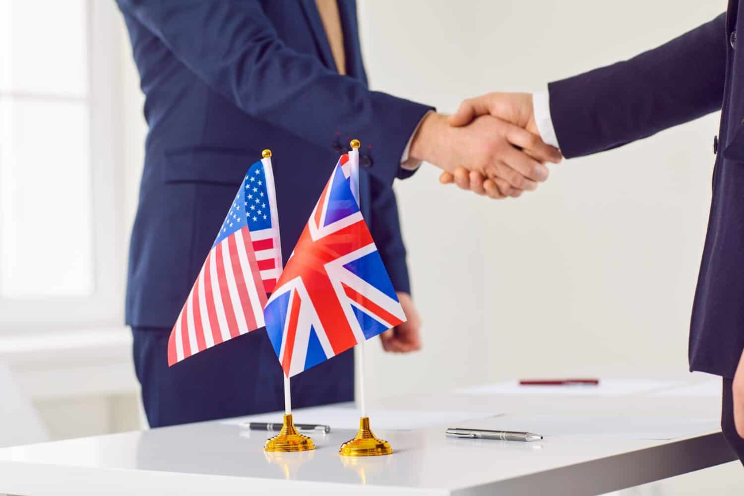 Diplomats from Britain and America meet together in office, reach bilateral trade agreement, sign business contract and exchange handshakes over negotiation table with national flags of USA and UK